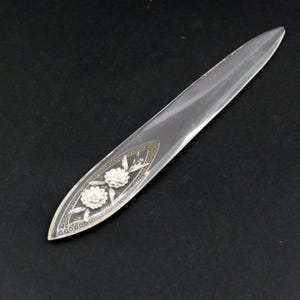 May include: A clear, elongated letter opener with a decorative floral design. The handle features two white flowers and a silver-colored border. The letter opener is made of a transparent material and has a pointed tip.