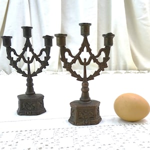 May include: Two antique bronze candle holders with ornate designs and three candle arms each. The candle holders are on a white lace tablecloth with a brown egg in the foreground.