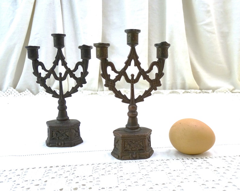 May include: Two antique bronze candle holders with ornate designs and three candle arms each. The candle holders are on a white lace tablecloth with a brown egg in the foreground.