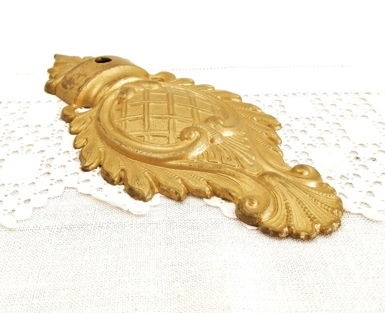 May include: A gold-toned metal decorative accent with a detailed, ornate design. The accent features a central, raised, geometric pattern surrounded by intricate scrollwork and leaf motifs.
