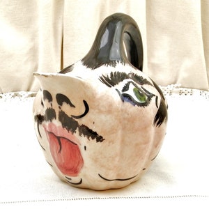 Puede incluir: Una jarra de cerámica con forma de cara con bigote negro, labios rojos y un ojo verde. La jarra está pintada en un estilo caprichoso y tiene un fondo blanco.