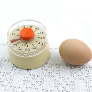 Peut inclure: Un minuteur de cuisine vintage avec un bouton orange et une base crème, à côté d'un œuf brun. Le minuteur a un couvercle en plastique transparent et un cadran numéroté. L'œuf est ovale et brun clair.