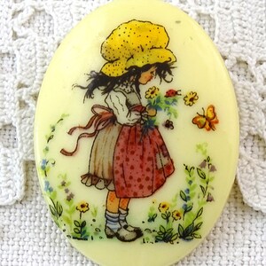 May include: A round, cream-colored button with a painted image of a girl in a yellow dotted hat holding a bouquet of flowers. The girl is wearing a white blouse and a red and white polka dot skirt. The background is a light green with flowers and butterflies.