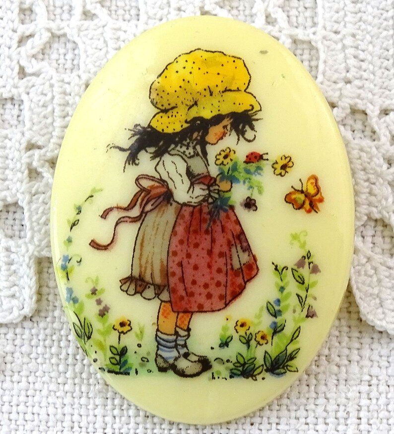 May include: A round, cream-colored button with a painted image of a girl in a yellow dotted hat holding a bouquet of flowers. The girl is wearing a white blouse and a red and white polka dot skirt. The background is a light green with flowers and butterflies.