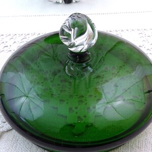 Puede incluir: Una tapa de vidrio verde con un pomo de vidrio transparente en la parte superior. La tapa es redonda y tiene un borde ligeramente elevado. El pomo tiene forma de espiral.
