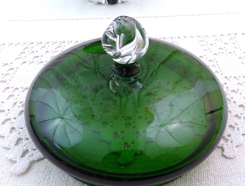 Puede incluir: Una tapa de vidrio verde con un pomo de vidrio transparente en la parte superior. La tapa es redonda y tiene un borde ligeramente elevado. El pomo tiene forma de espiral.