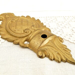 May include: A gold-colored metal decorative ornament with intricate floral and scrollwork designs. The ornament has a central circular design with a grid pattern and a hole in the center.