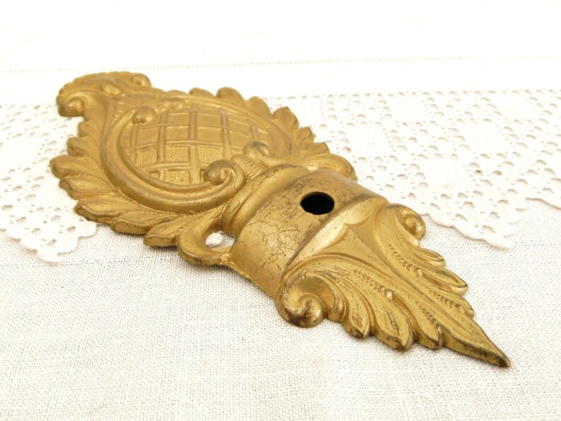 May include: A gold-colored metal decorative ornament with intricate floral and scrollwork designs. The ornament has a central circular design with a grid pattern and a hole in the center.