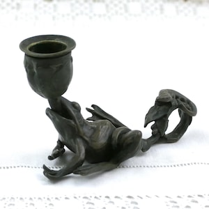 May include: A dark bronze-toned metal candle holder shaped like a frog. The frog is in a seated position with its mouth open, supporting a cup-shaped candle receptacle. A decorative handle with leaf-like details extends from the frog's back.