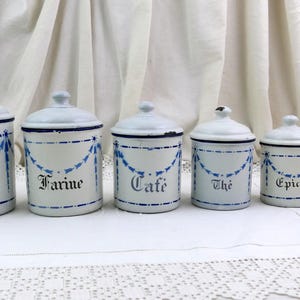 Könnte beinhalten: Ein Set aus sechs weißen, emaillierten Metalldosen mit Deckeln, verziert mit blauen Girlanden und schwarzen Beschriftungen: Sucre, Farine, Cafe, The, Epices und Poivre. Die zylindrischen Behälter sind nach ihrer Größe absteigend angeordnet.