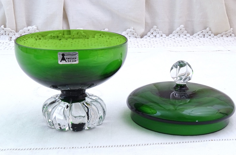Puede incluir: Un cuenco de caramelo de vidrio verde con una tapa de vidrio transparente y una perilla de vidrio transparente. El cuenco tiene una base de vidrio transparente con un dise&ntilde;o festoneado. El cuenco tiene una etiqueta que dice "Sweden Aseda".