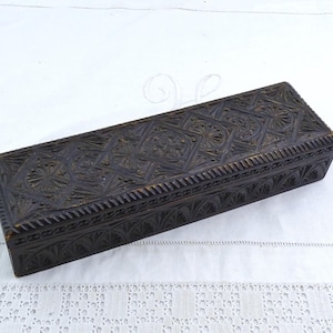 Puede incluir: Una caja rectangular de madera marrón oscuro con intrincados patrones tallados geométricos y florales en la tapa y los lados. La caja se encuentra sobre un fondo blanco texturizado, mostrando su detallada artesanía.