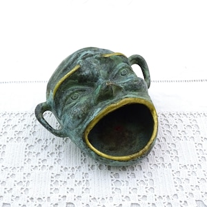 May include: A green and gold metal ashtray shaped like a face with an open mouth. The ashtray is on a white crocheted surface.