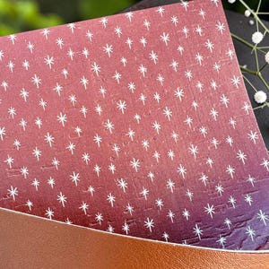 May include: A rectangular sheet of dark maroon material featuring a repeating pattern of small white starburst icons, partially tucked into a smooth, tan-colored leather-like sleeve with a curved opening edge.