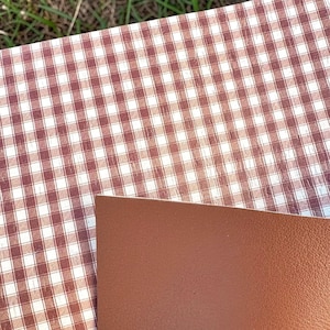 May include: A sheet of material featuring a brown and white gingham check pattern on one side and a solid, smooth, matte brown surface on the reverse side. The material is flexible and displays a consistent, repeating grid design.