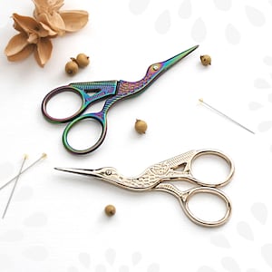 May include: Two pairs of decorative scissors with bird-shaped handles. One pair is iridescent and the other is gold. Both pairs are on a white surface with small brown objects and two sewing pins.