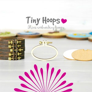 Puede incluir: Un conjunto de cuatro aros de bordado de madera, con una base de madera marrón y tornillos de metal dorado. Los aros están apilados uno encima del otro, con el aro más pequeño en la parte superior. Los aros están etiquetados como "Tiny Hoops Mini embroidery hoops".