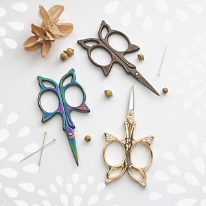 May include: Three pairs of butterfly-shaped scissors with different colored finishes: iridescent, bronze, and gold. The scissors are on a white background with a decorative pattern.
