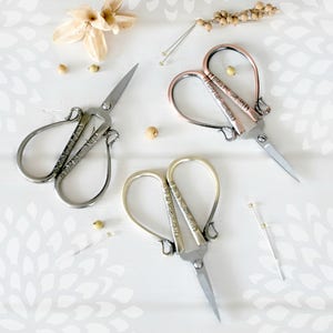 May include: Three pairs of decorative heart-shaped scissors with ornate designs. The scissors are silver, gold, and rose gold.