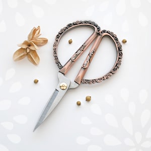 May include: A pair of ornate, copper-toned scissors with a silver blade. The scissors have a decorative design with floral motifs.