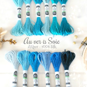 May include: An assortment of blue and teal silk embroidery floss skeins. The image features various shades of blue, from light to dark, with the text "Au ver à Soie d'Alger - 100% Silk". The floss is arranged in a visually appealing display.