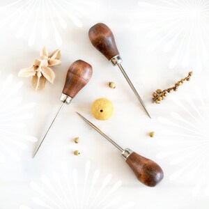 May include: Three wooden handled tools with metal tips, used for leather crafting. The tools are on a white surface with small yellow and brown objects scattered around them.