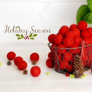 以下が含まれることがあります： 鮮やかな赤いフェルトボールがいっぱい入ったワイヤーバスケット。小さなベルと松ぼっくりが散らばっています。「Holiday Season」の文字がバスケットの上に書かれています。背景には緑の葉があります。