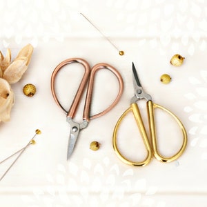 May include: Two pairs of small scissors on a white surface. One pair is rose gold and the other is gold.