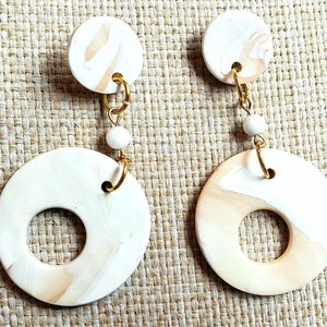 May include: A pair of cream-coloured dangle earrings. Each earring features a small circular stud, a small white bead, and a large circular pendant with a hole in the centre. The earrings are connected with gold-coloured wire.