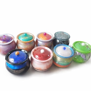 Pode incluir: Uma coleção de pequenos potes de vidro decorativos com tampas. Os potes são de várias cores, incluindo roxo, azul, verde, laranja e rosa. Cada pote tem um design e esquema de cores únicos, com um acabamento brilhante.