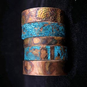 May include: A stack of five metal cuff bracelets. The top band has a hammered copper texture. The second and fourth bands feature a bright turquoise patina finish. The third band is smooth copper with a small stamp, and the bottom band is smooth copper.