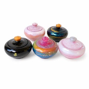 Pode incluir: Cinco potes redondos decorativos com tampa em várias cores. Os potes incluem preto com um detalhe dourado, rosa com redemoinhos brancos, um design de arco-íris e um azul escuro com detalhes dourados. Cada pote tem um pequeno botão na tampa.