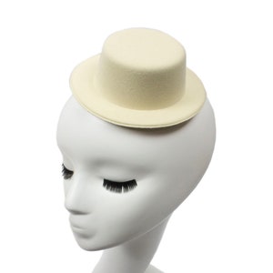 Puede incluir: Un pequeño sombrero de copa de fieltro de color blanco roto con un ala ancha. El sombrero está colocado sobre una cabeza de maniquí blanca.