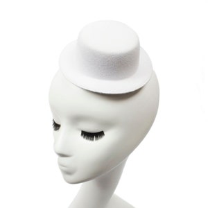 Könnte beinhalten: Ein kleiner, weißer Zylinder sitzt auf einem weißen Schaufensterkopf mit geschlossenen Augen und langen, schwarzen Wimpern. Der Hut ist zylindrisch mit flachem Oberteil und Krempe. Die Schaufensterpuppe ist glatt und minimalistisch.