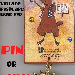 May include: Vintage postcard featuring a cartoon groundhog with the text "Ground Hog Day" and "Feb. 2". The postcard is attached to a gold sun charm with a face.