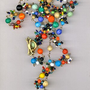 May include: A colorful beaded necklace with a vintage illustration of a woman in a hat. The necklace is made with a variety of glass beads, including clear, red, blue, green, and yellow beads. The illustration is framed with metal and decorated with small metal flowers.