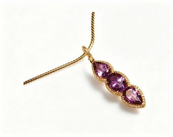 May include: A gold pendant necklace featuring three teardrop-shaped purple gemstones. The gemstones are set in a gold frame with small, clear accent stones. The necklace has a delicate gold chain.