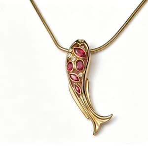 May include: A gold-toned necklace featuring a fish-shaped pendant. The pendant is decorated with multiple marquise and oval-cut red stones and small clear round stones set within the gold metal body, suspended from a thin gold-toned snake chain.