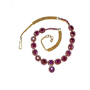 May include: A gold-toned necklace with a series of deep red gemstones. The necklace features a central pendant and a matching bracelet. The gemstones are set in a floral design, creating a delicate and elegant look. The necklace is displayed on a white background.