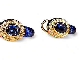May include: A pair of gold-colored cufflinks with a gold filigree design around a dark blue oval stone. The cufflinks have a curved post for securing to a shirt cuff. The background is white.