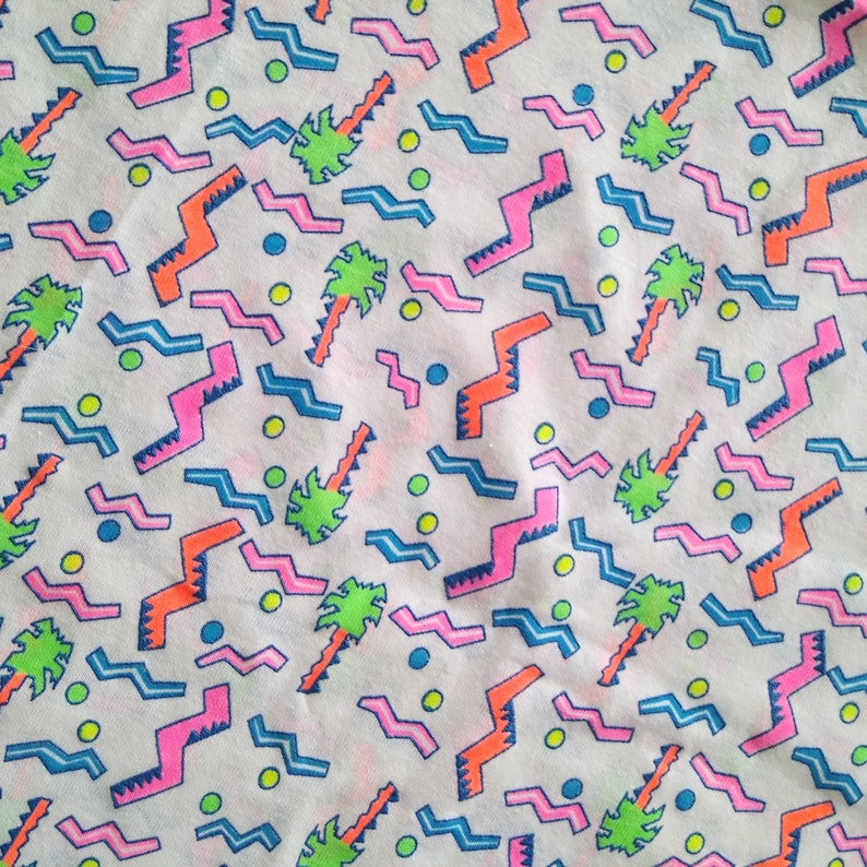 May include: A white fabric with a repeating pattern of pink, blue, and orange geometric shapes and green palm trees. The background is white with small yellow and blue dots.