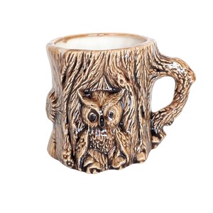 May include: A ceramic coffee mug shaped like a tree stump, featuring a detailed relief of an owl. The mug is primarily brown with lighter brown and beige accents, and has a textured surface. The handle is designed to look like a tree branch.