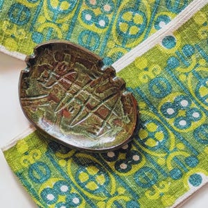 May include: An oval-shaped ceramic dish with a glossy, mottled brown and green glaze featuring an incised geometric pattern. The dish is placed on a textured fabric surface with a repeating green and teal circular and linear print pattern.