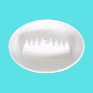May include: A round, white ceramic ashtray featuring a smooth, glossy finish. The interior of the basin includes a series of integrated notches along one side, designed to hold items securely in place within the circular, shallow bowl structure.