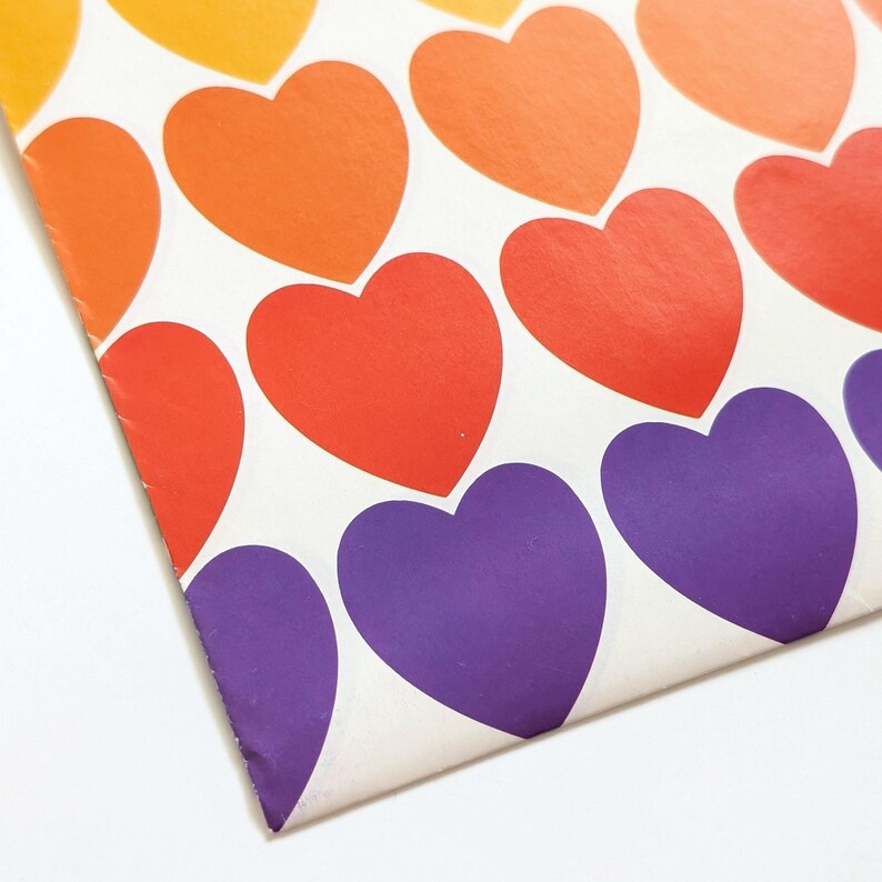 Pu&ograve; includere: Primo piano di una superficie bianca con un motivo di cuori colorati. I cuori sono in tonalit&agrave; arancioni, rosse e viola. La carta presenta una leggera piega nell'angolo.
