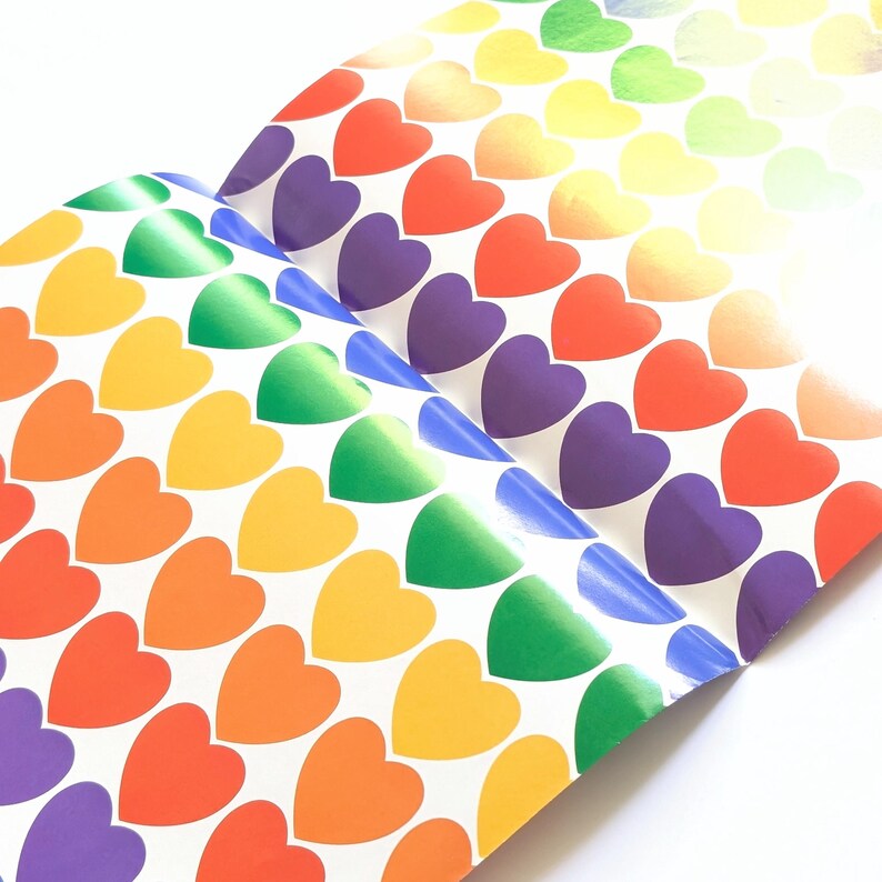 Pu&ograve; includere: Fogli di adesivi a forma di cuore arcobaleno. I cuori passano attraverso i colori dell'arcobaleno, dal rosso all'arancione, giallo, verde, blu e viola. Gli adesivi sono disposti in file e colonne su sfondo bianco.