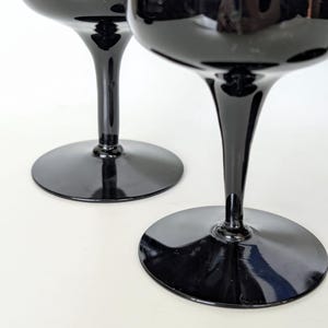 May include: Two black glass wine glasses with a tall stem and a wide bowl. The glasses are on a white surface.