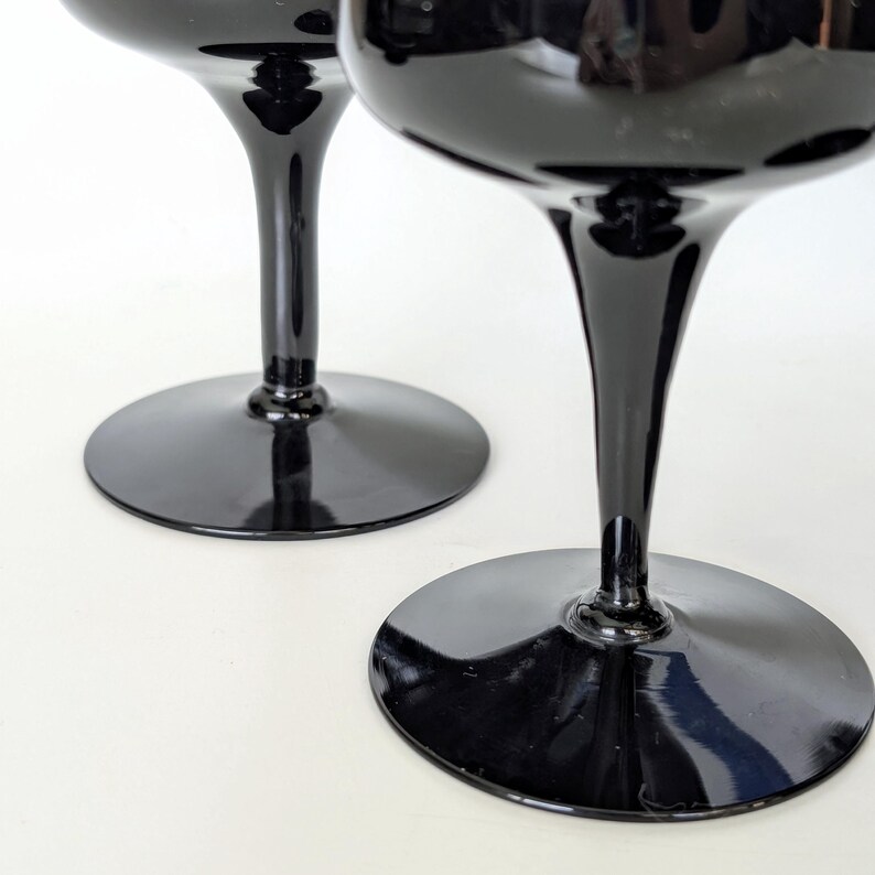 May include: Two black glass wine glasses with a tall stem and a wide bowl. The glasses are on a white surface.