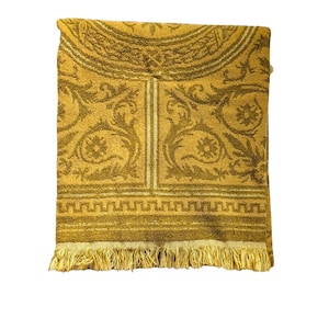 May include: Mustard yellow bath towel with an ornate, repeating floral and geometric pattern. The towel has a decorative border and a fringe along the bottom edge. The design is in a darker shade of yellow, creating a textured appearance.
