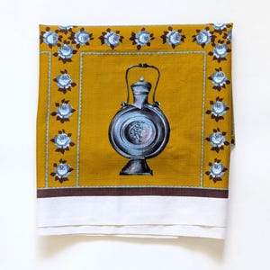 May include: Mustard yellow kitchen towel with a central illustration of a silver flask. The towel is bordered with blue roses and a brown trim. The bottom edge is white.